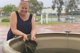 A woman stands over a large tub filled with water, holding a potted seagrass plant, mud and trees are visible behind.