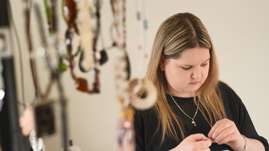 Keely Payne sits at her dining table making jewellery, with hanging necklaces in the foreground.