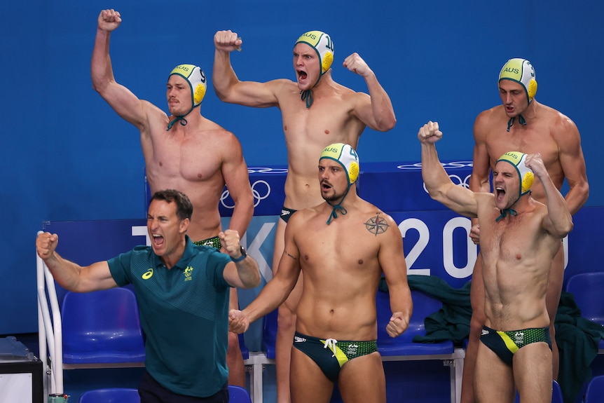 A group of men in green and gold speedos raise their hands in the air and shout in celebration