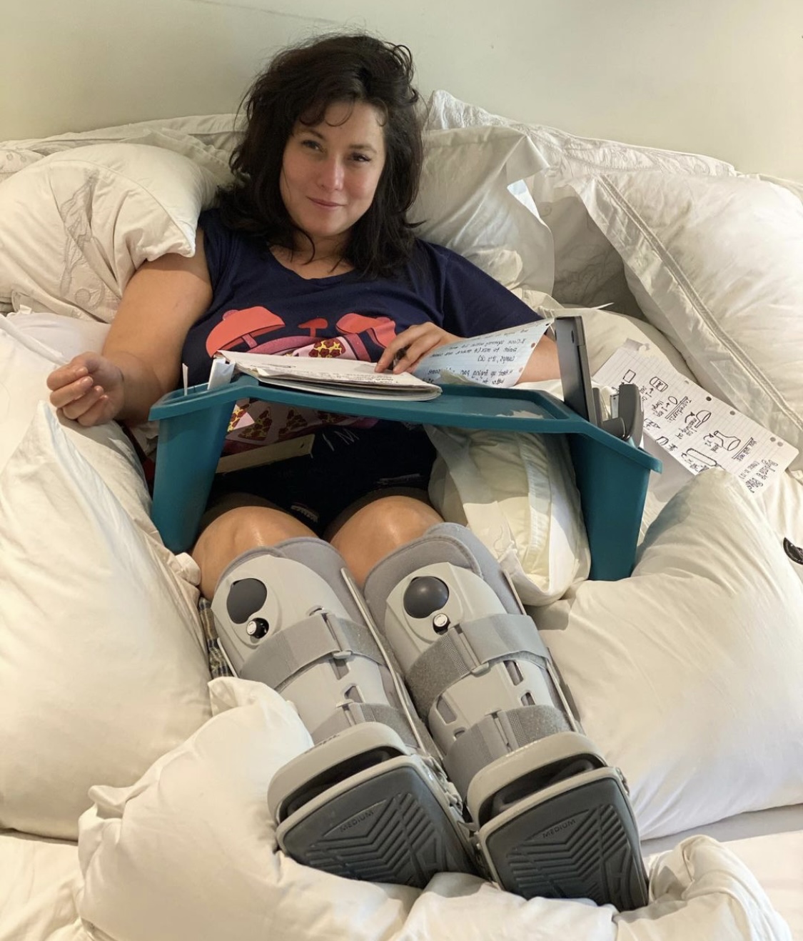 Elly-May in a hospital bed writing, wearing boots on both legs.