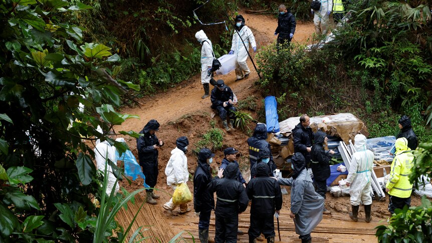 Workers wearing full PPE suits carry boxes and bags down a winding dirt trail. 
