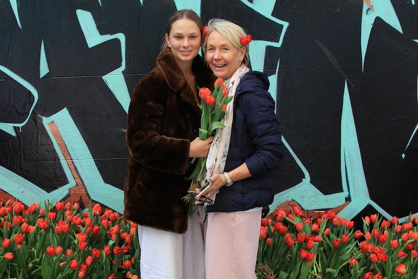 Charlotte and her mum Ann McArdle stand in front of a row of orange tulips with a graffiti wall behind them.