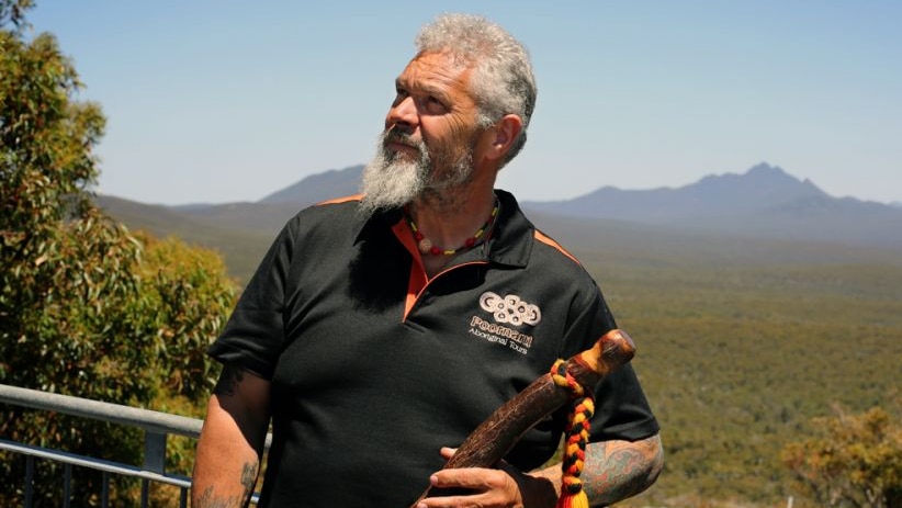 An Indigenous man stands amongst bushland with mountain range in background.