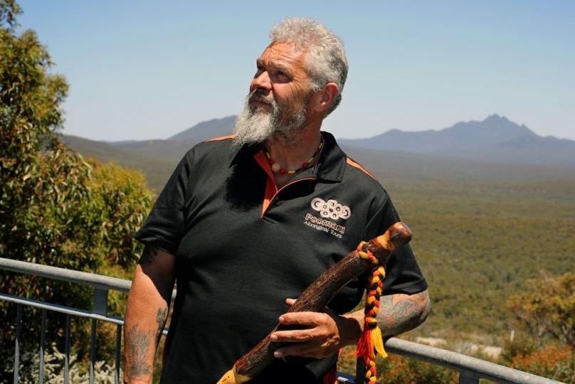 An Indigenous man stands amongst bushland with mountain range in background.