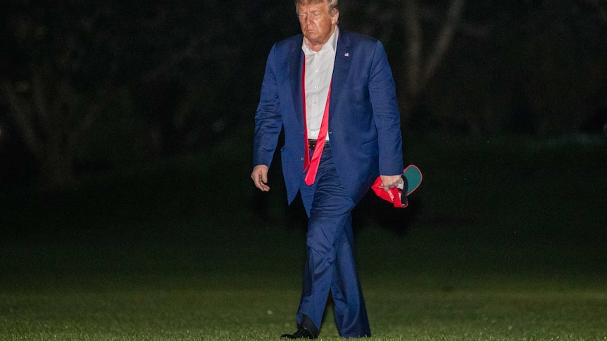 Trump walks on the lawn with his shirt unbuttoned, tie undone and looking down towards the ground