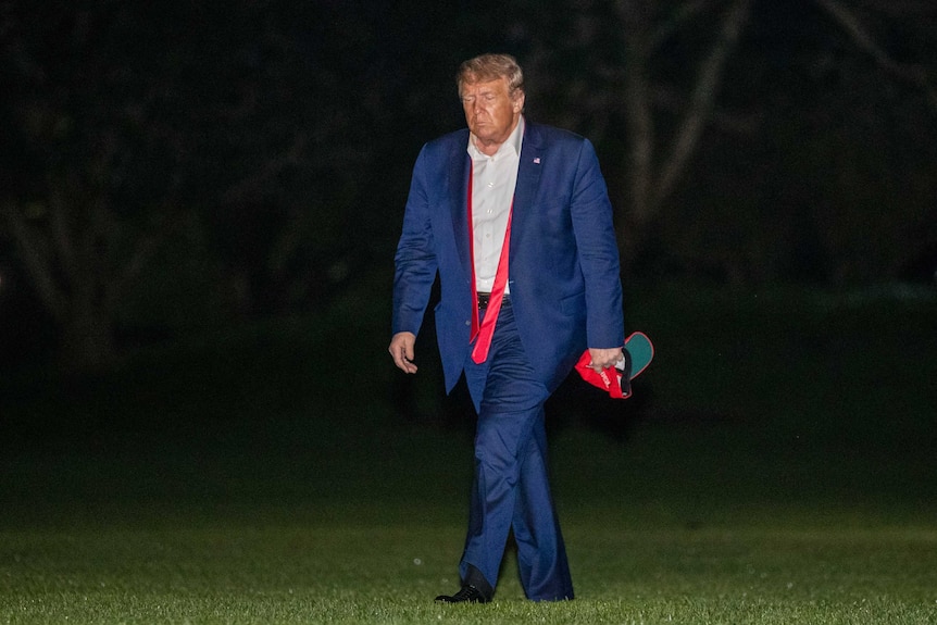 Donald Trump looking sad while walking across a lawn at night with his tie untied and a MAGA hat in his hand