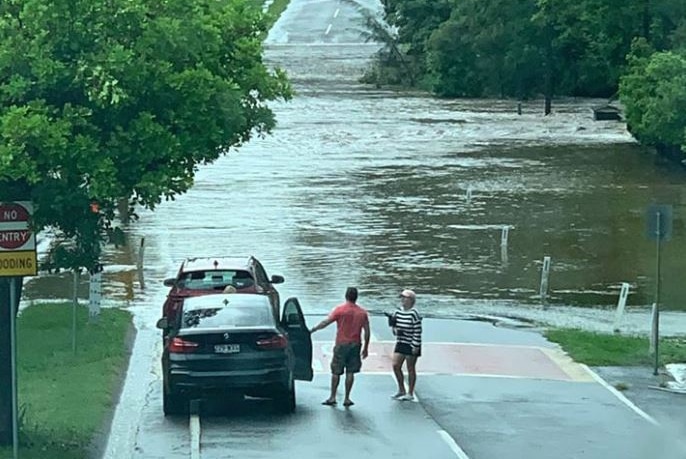 Two people stand outside two cars before water on a flooded road with trees around them