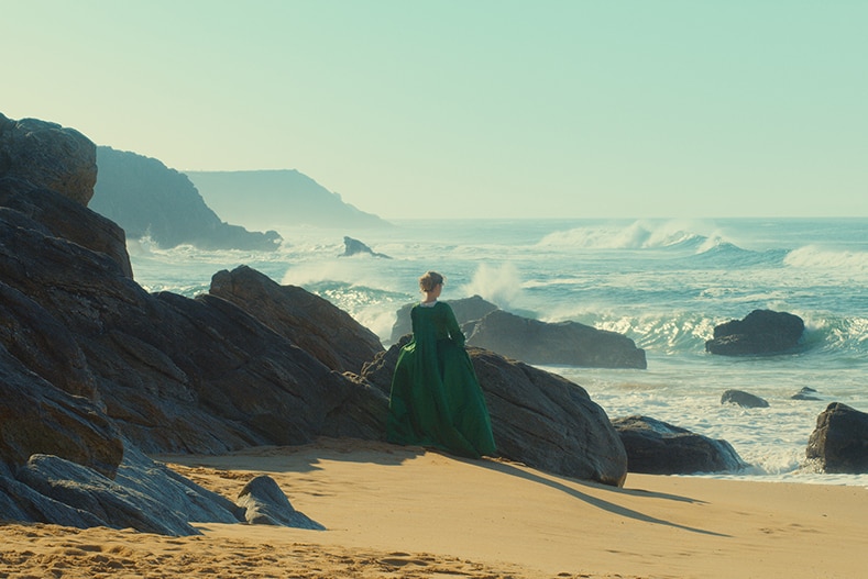 A woman wearing a green 18th century period dress stands alone near rocks on beach, looking out towards rough ocean.