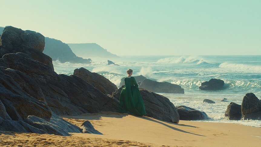 A woman wearing a green 18th century period dress stands alone near rocks on beach, looking out towards rough ocean.