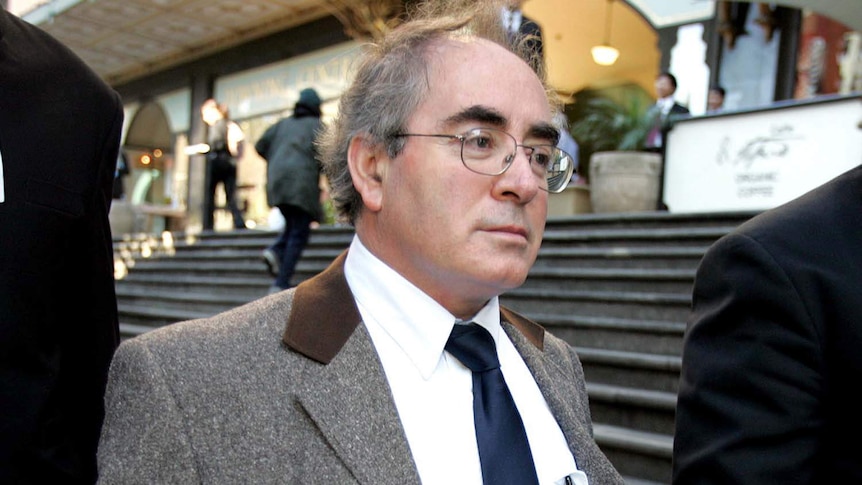 A bespectacled, balding man in a tweed suit leaves a court building.
