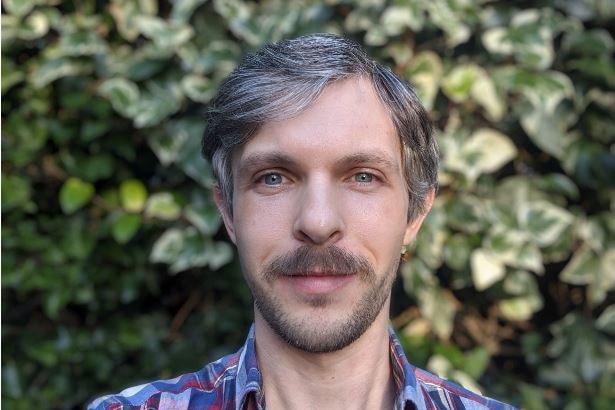 A man with a trimmed beard stands in front of a hedge, he is wearing a plaid shirt.