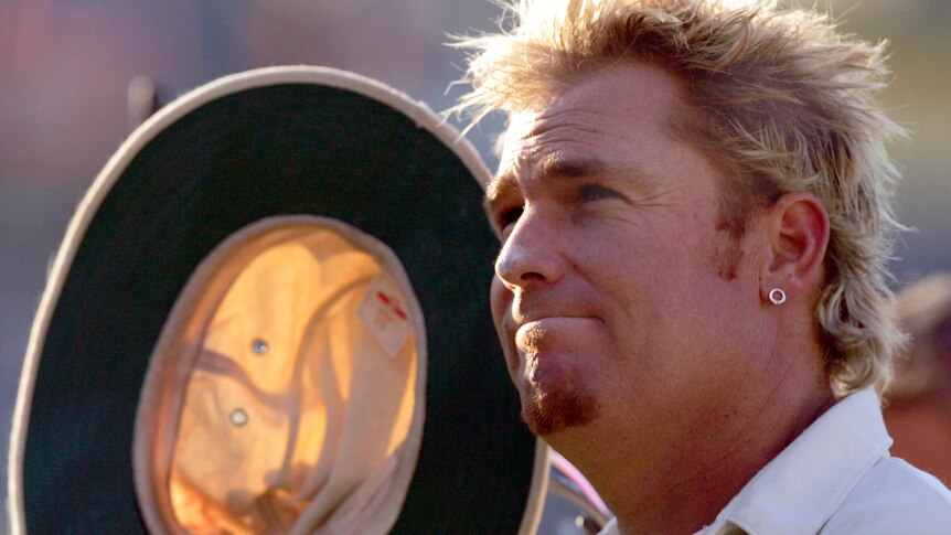 Close-up of Shane Warne holding his hat up beside his face