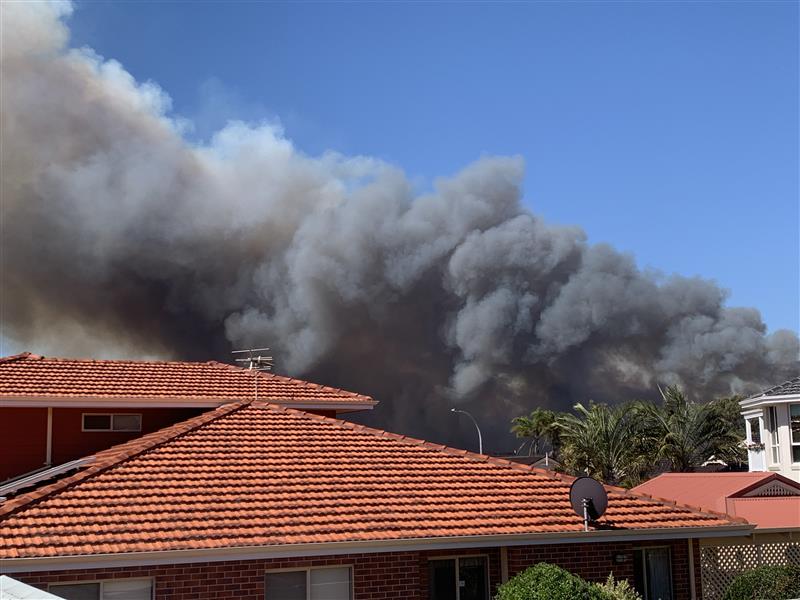 En primer plano, una casa con una gran nube de humo negro que se eleva sobre el horizonte.