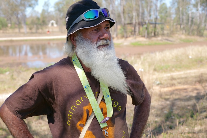 Milton Lawton standing with a brown Carnarvon Gorge t-shirt on, hands on hips, grass and water behind.
