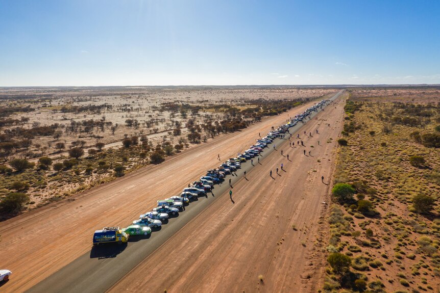 An aerial shot of a straight, dusty road in a desert with a line of cars travelling along it.