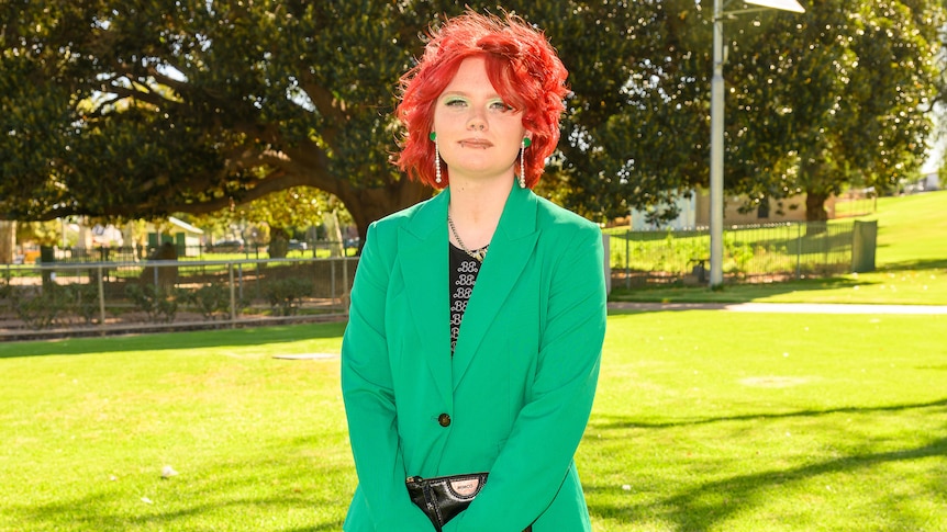 A young person with white skin and red hair wearing a green suit stares at the camera in a park. 