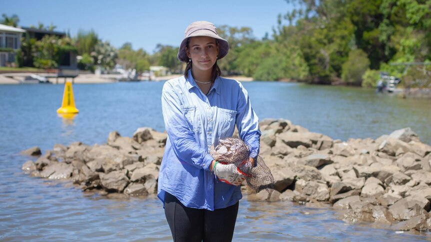 A woman stands beside a water source holding a mesh bag of shellfish