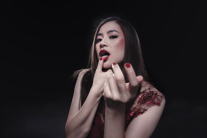 An Asian Australian woman with red lips and rouged cheeks looks seductively toward the camera with one hand outstretched.