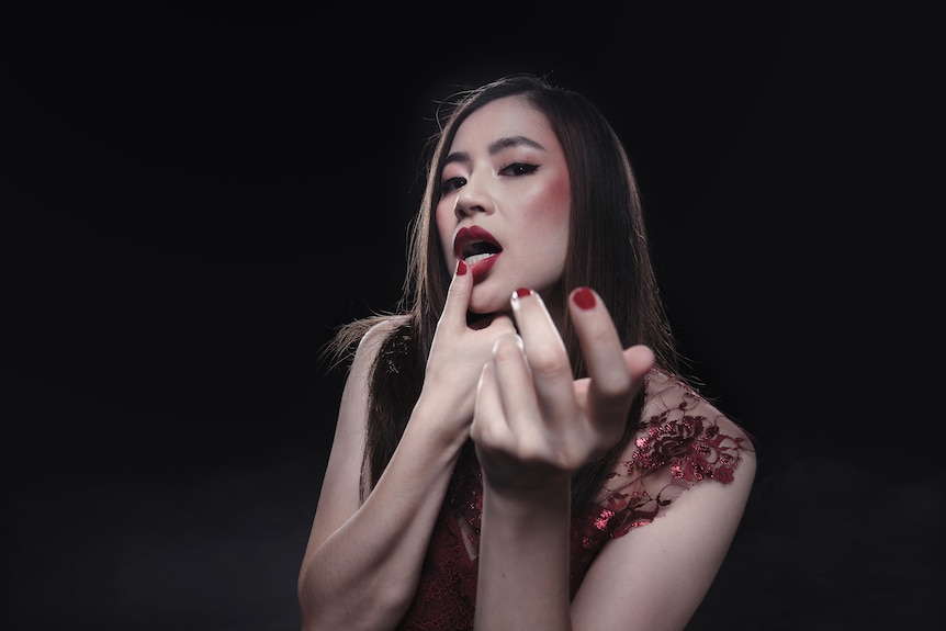 An Asian Australian woman with red lips and rouged cheeks looks seductively toward the camera with one hand outstretched.