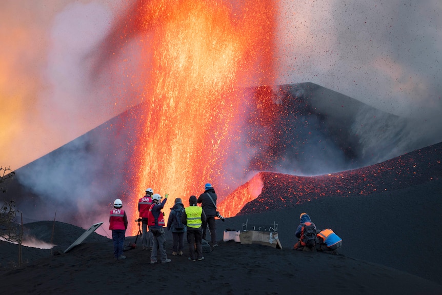 A wave of lava flies into the air as people in high-vis vests look on.