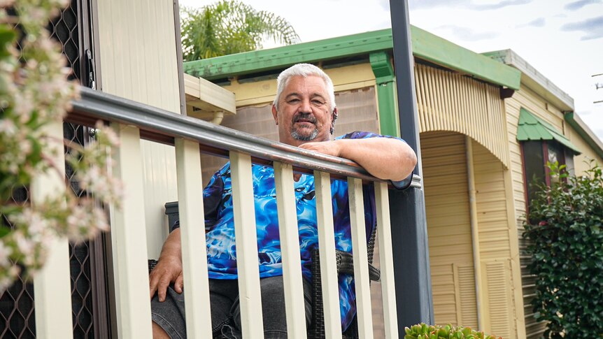 An older man with white hair and a neat beard sits out the front of his house.