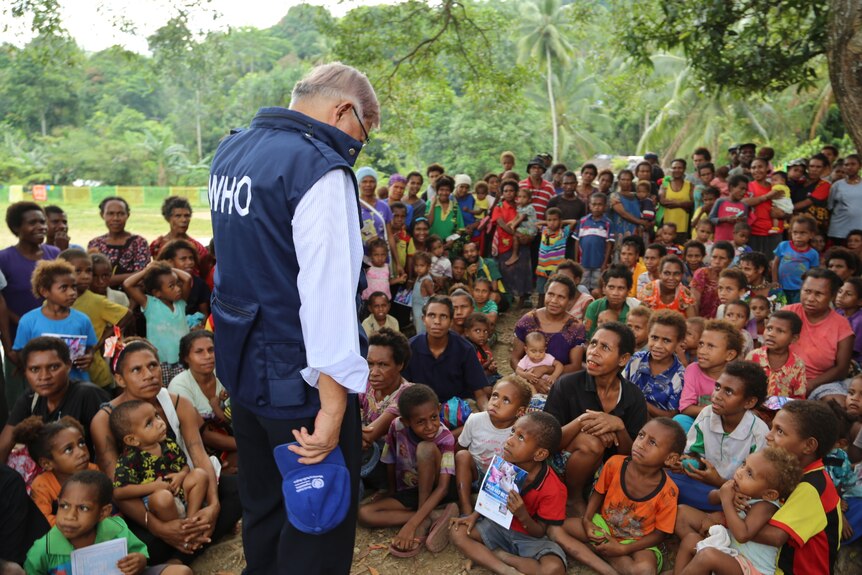 A doctor speaking to a crowd of people, many of whom are sitting on the ground.