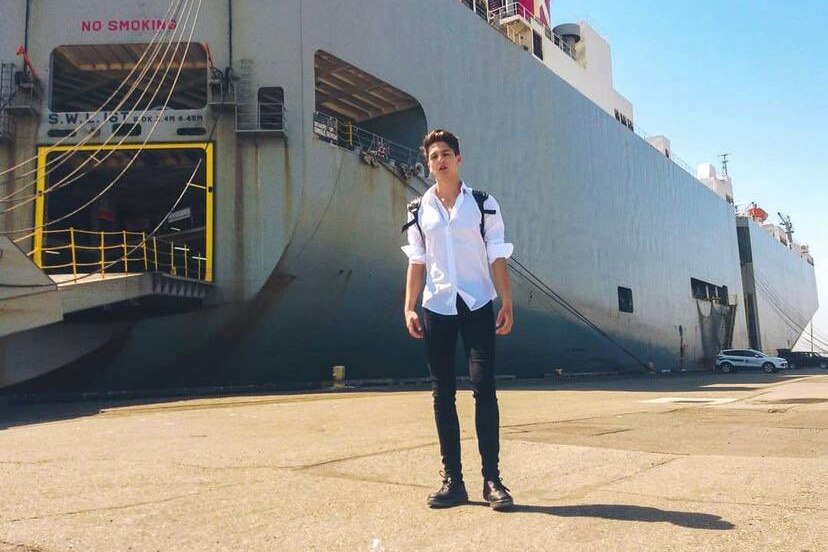 He stands in a white shirt and black pants near a huge ship