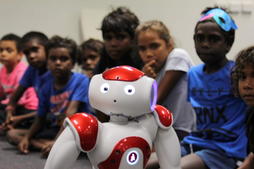 A robot looks up at the camera while students behind it look at the robot.