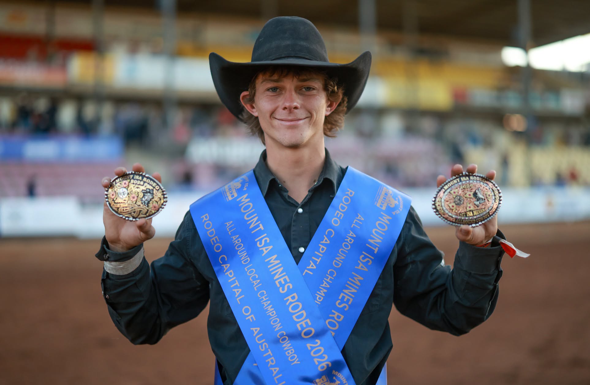A young cowboy in a black hat and blue championship sashes holds two rodeo buckles in the arena.