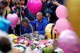 The stepfather of a victim of the Manchester attack mourns in front of flowers and balloons, surrounded by people.