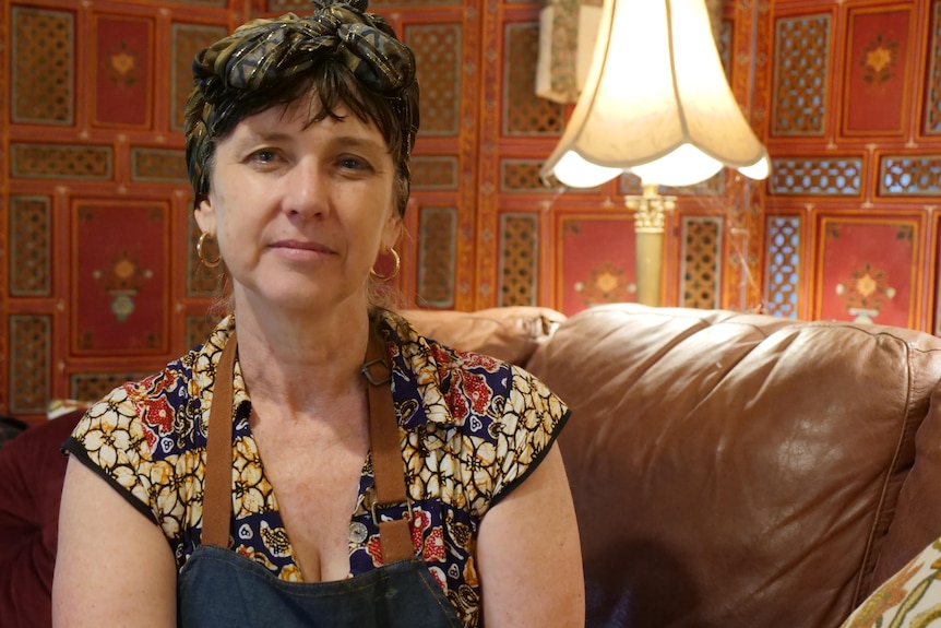 A woman wearing a hair scarf and blue apron sits on a lounge in front of a decorative screen.