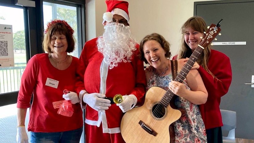 Jo Fergusn-Allen stands holding a guitar in a group photo with her daughter Jen, a choir member and Santa. Everyone is laughing.