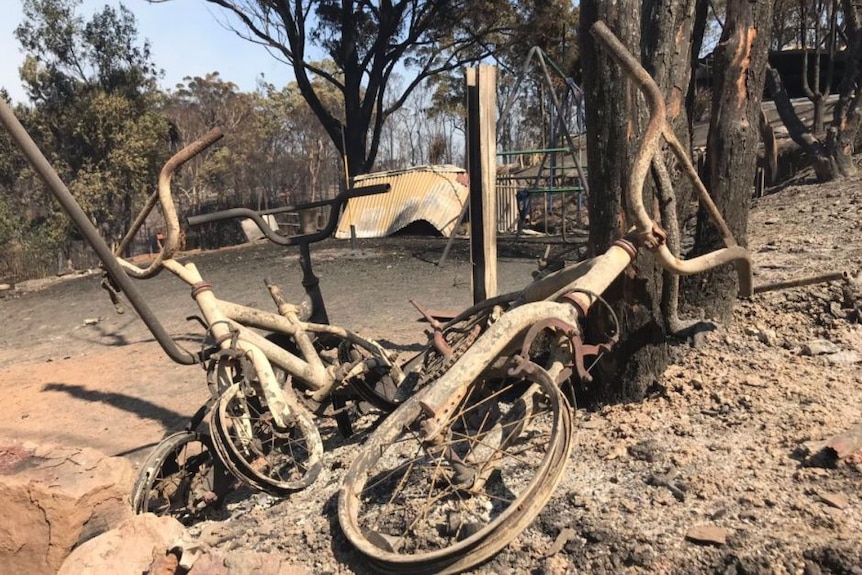 Two grey and brown burnt bicycles in the foreground in the town of Tathra. Ash, trees and burnt playground in the background.