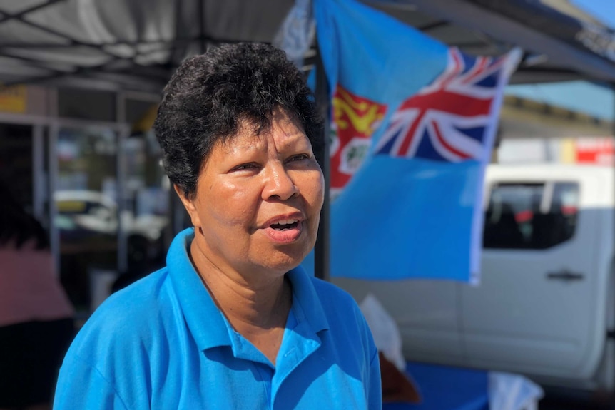 Aunty Violet Langan looks slightly away from the camera as she stands mid-speech in front of the Fiji flag.