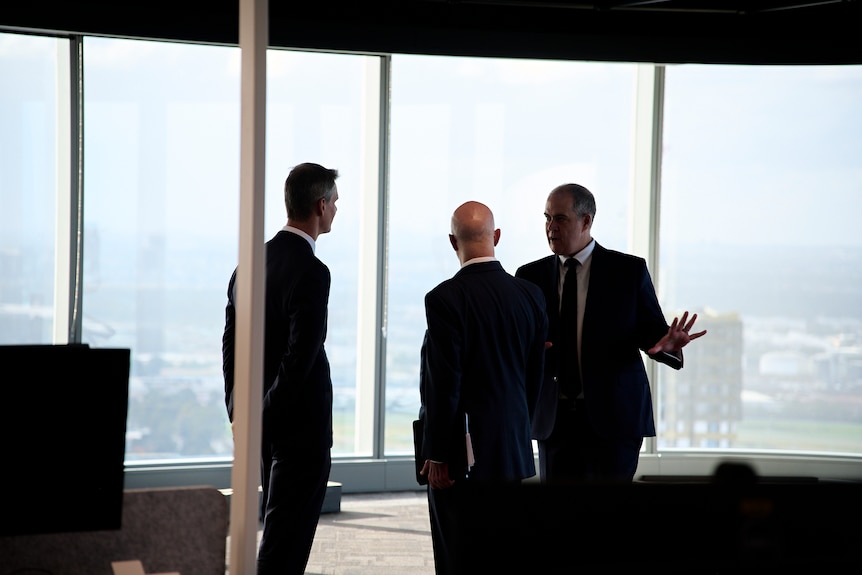 Three men in suits talk to each other standing in front of big windows. 