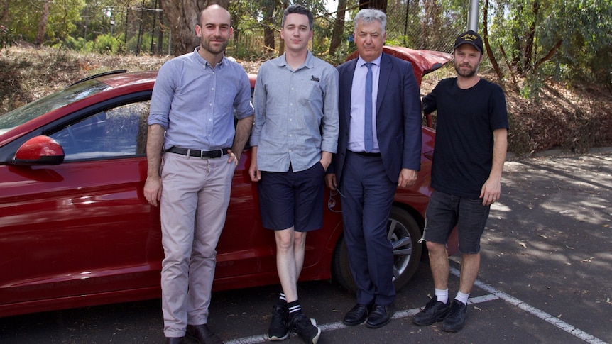 Four men stand in front of a red car.