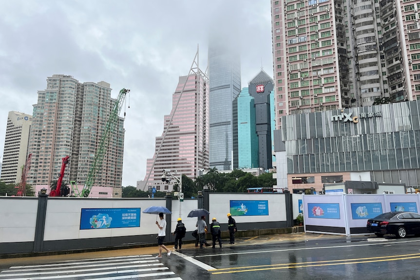 Four workers drain water from a flooded construction site as a person walks past with high rise buildings in the distance.