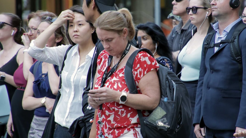 Men and women stand looking ahead, waiting to cross the road. Some have headphones others are looking at their phone.