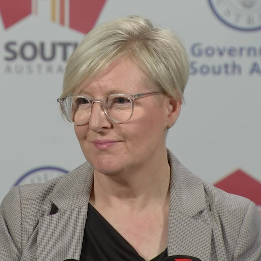 A woman with short blonde hair, clear glasses and a grey blazer smiles looking off camera in front of an SA government banner
