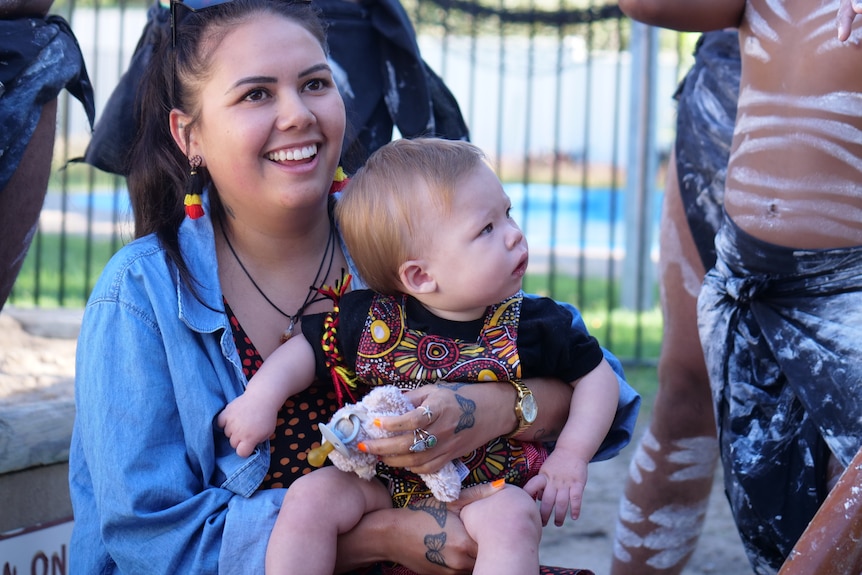Baby Jarli, black shirt, Aboriginal dot painted jumpsuit, woman in blue shirt holding him smiling.