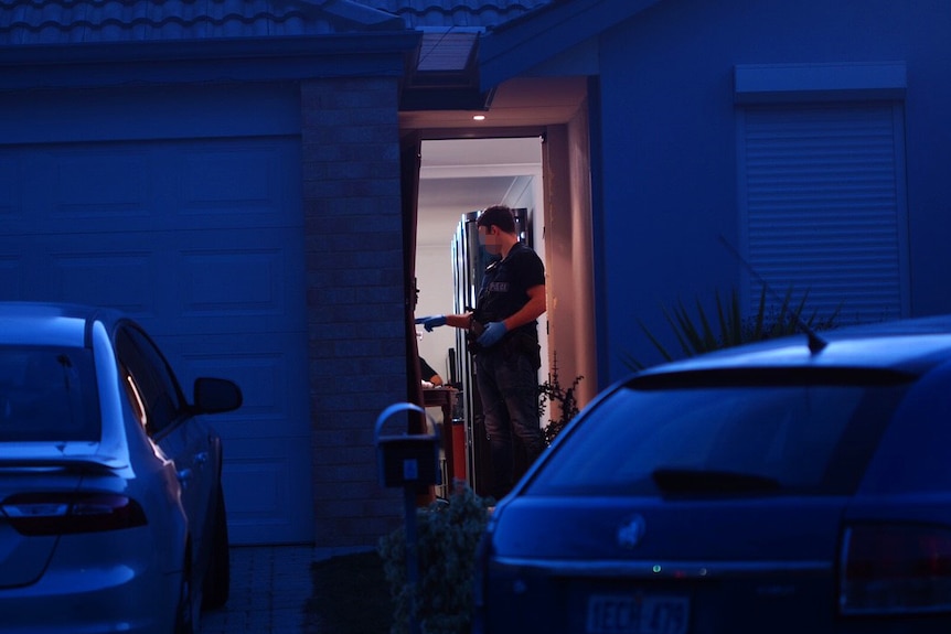 A policeman in the doorway of a house conducting a dawn drugs raid, with cars in the foreground.