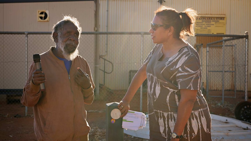Joseph Lane hold a microphone and talks to Leanne Liddle in front of a fence in Haasts Bluff.