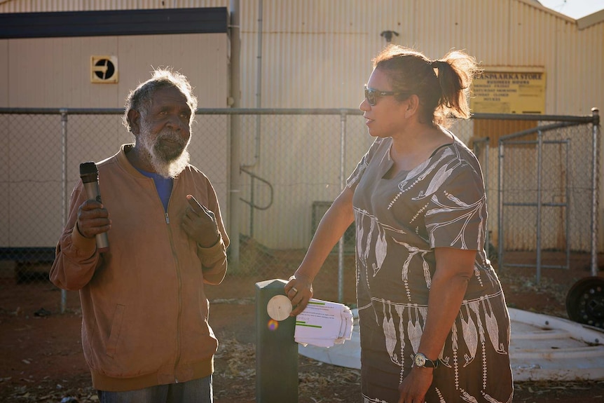 Joseph Lane hold a microphone and talks to Leanne Liddle in front of a fence in Haasts Bluff.