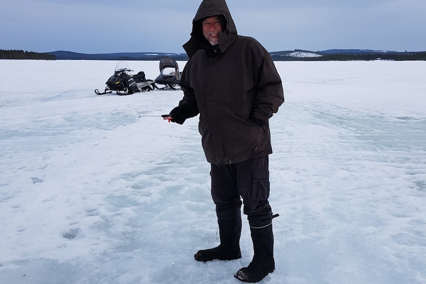 A man in heavy waterproofs stands in an icey landscape, snowmobile behind.