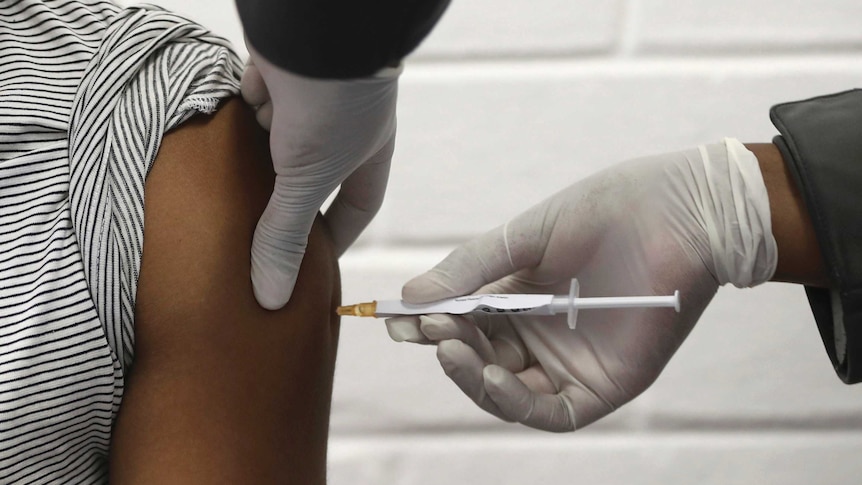 The shoulder of a volunteer is seen as they receive an injection by a person wearing gloves.