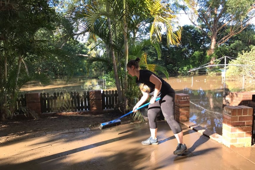 A woman named Caroline sweeps mud from the back of her home which is surrounded by flooding