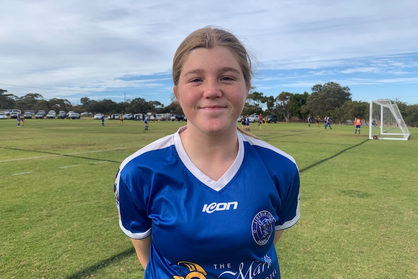 A 14-year-old girl with blonde hair smiling in front of a soccer field in a blue soccer jersey.