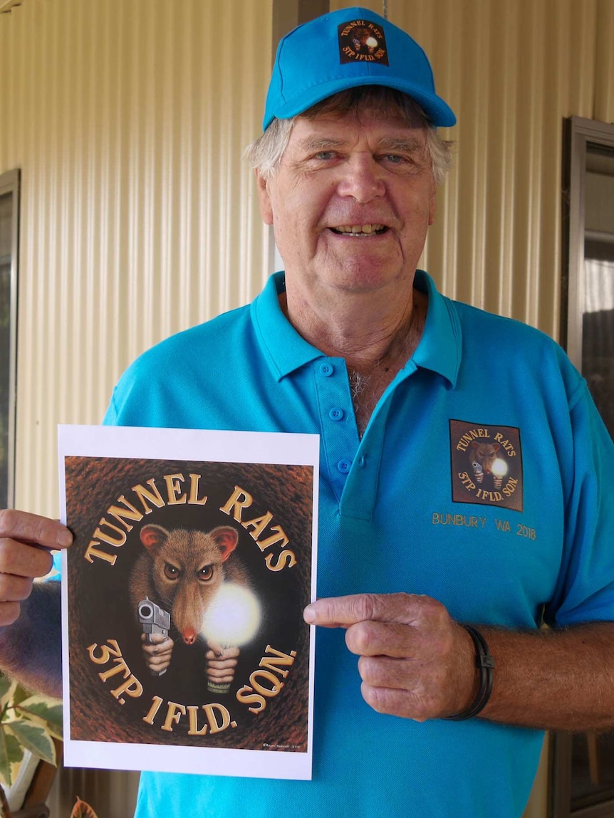 A man with a blue shirt and hat standing in front of a metal panel house holding an logo saying Tunnel Rat 3TP. 1FLD. SON.