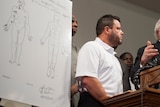 Shawn Parcells stands at a lectern during a news conference. There is a diagram of a body presented next to him. 