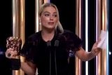 Margot Robbie holds a BAFTA statue while speaking at a microphone with her hands raised.